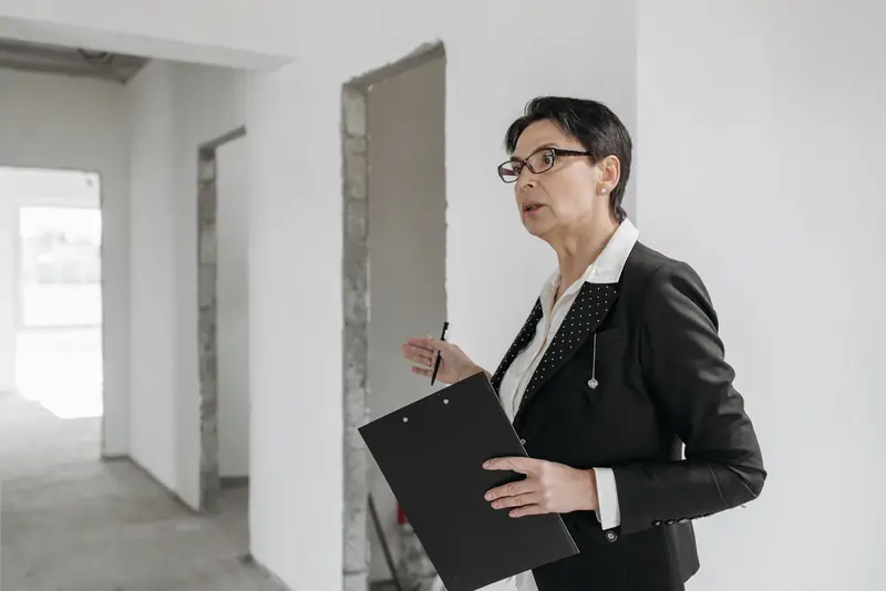 Woman in a dark suit holding a clipboard while inspecting an unfinished interior with bare walls and a concrete floor