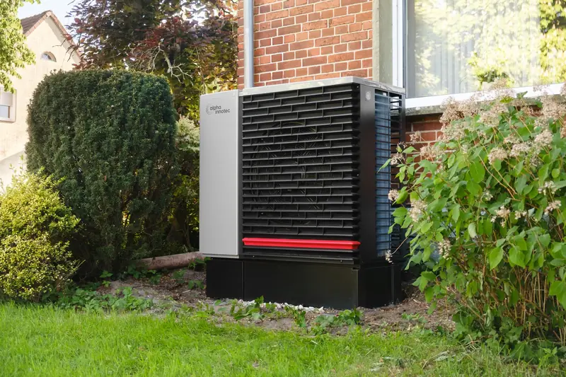 Air source heat pump unit standing on a base in the front garden of a red-brick house, between a clipped hedge and a flowering shrub