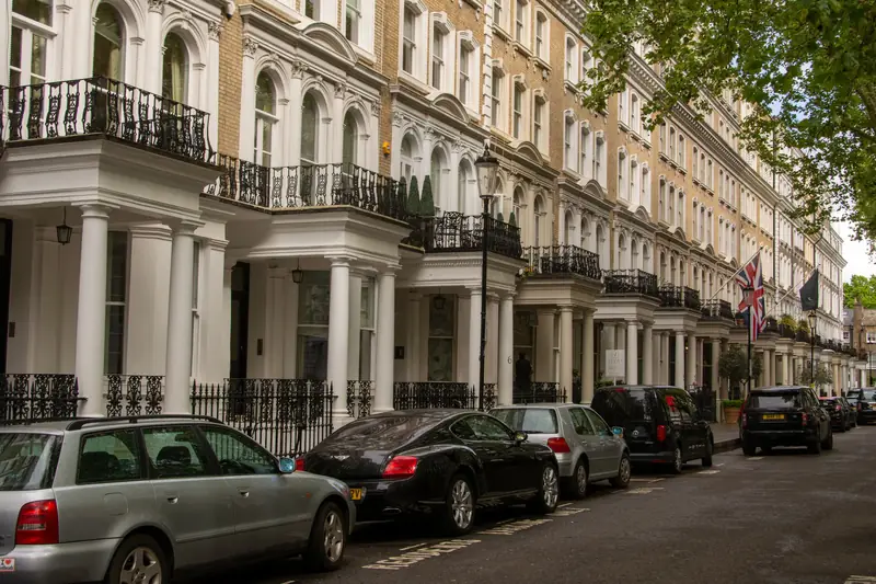 A long row of white stucco Victorian terrace houses in London with pillared porches, iron balconies and tall sash windows, with cars parked along the street
