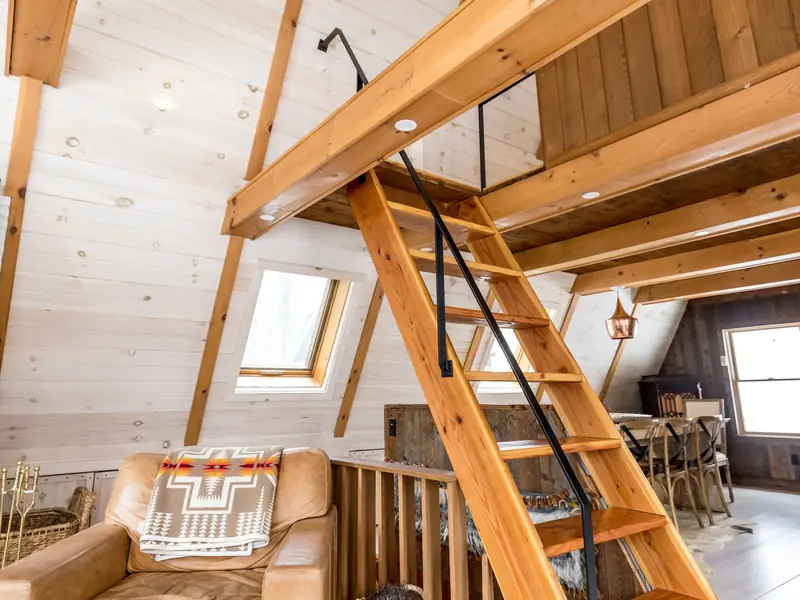 Steep timber stair with a black metal handrail rising to a mezzanine floor in a bright attic room with pale timber-clad walls, exposed beams and small gable windows