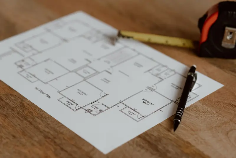 Printed first-floor plan drawing on a wooden desk, with a pen and a tape measure beside it