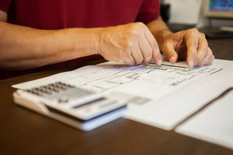 A person at a wooden desk checking a printed house floor plan with a ruler, with a desk calculator in the foreground