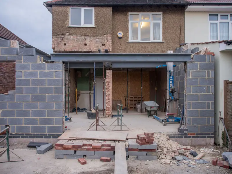 Single-storey rear extension under construction on a semi-detached house, with blockwork side walls, a steel beam across the opened-up rear wall and bricks stacked in the garden